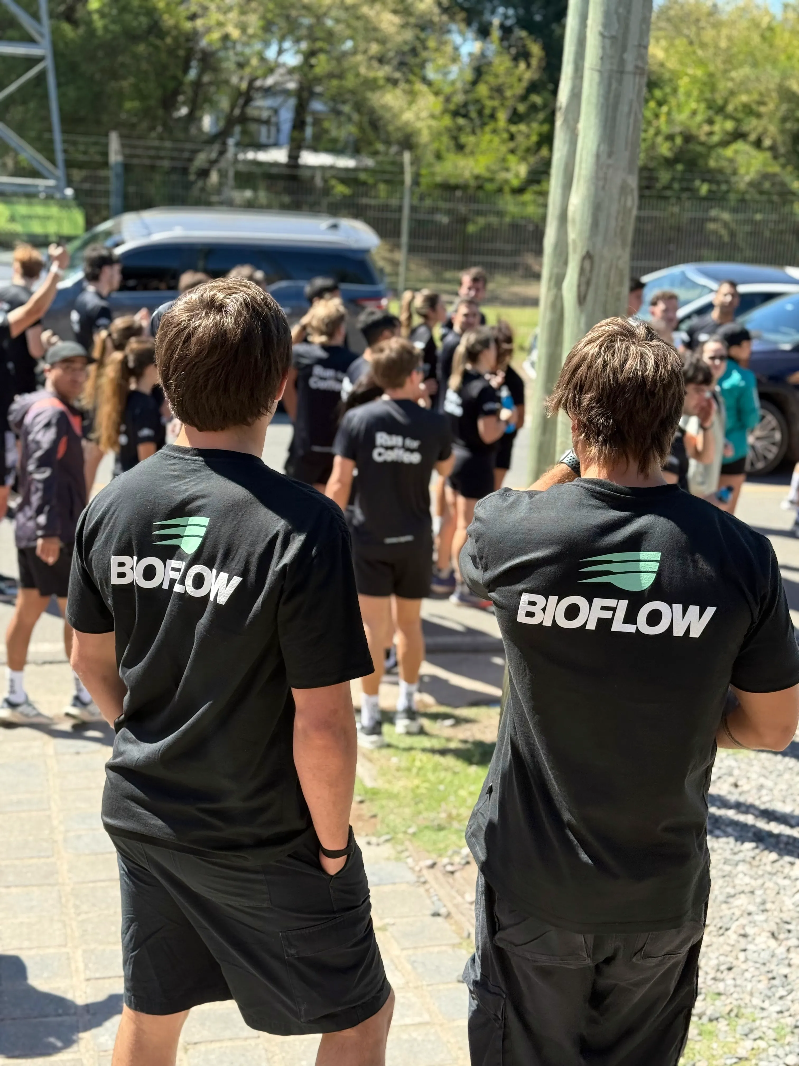 Dos personas de espaldas con remeras de Bioflow, en la previa de una carrera con corredores reunidos.