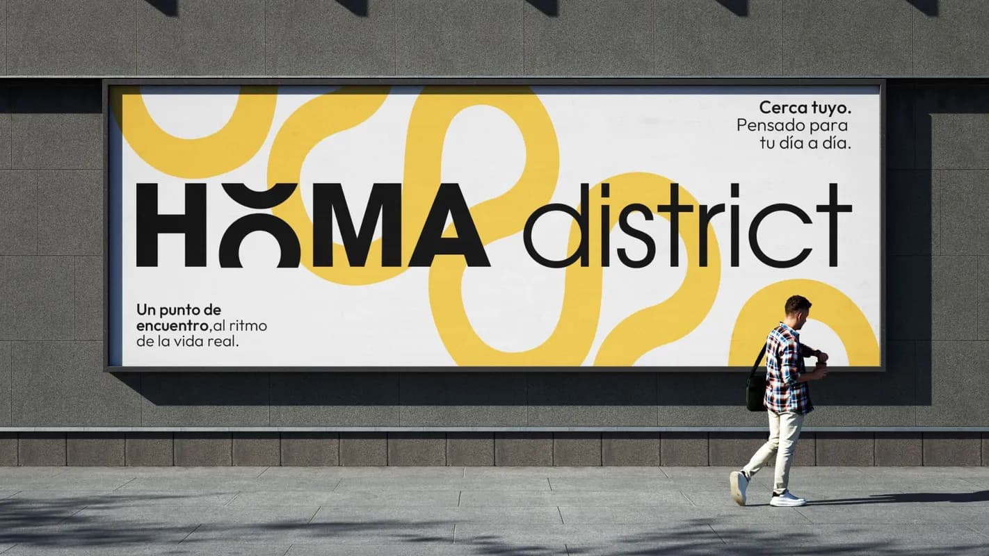 Cartel de HOMA District en una pared de la calle, con curvas amarillas sobre fondo blanco y la frase: un punto de encuentro, al ritmo de la vida real.